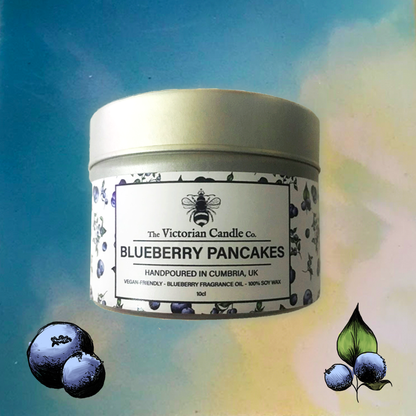 A 100ml tin of The Victorian Candle Co.'s Blueberry Pancakes Scented Candle is showcased against a sky-like background. This vegan-friendly home fragrance features illustrations of blueberries, enhancing its fruity appeal.