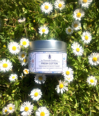 The Fresh Cotton - Scented Candle (100ml Tin) from The Victorian Candle Co. with 100% soy vegan-friendly wax is nestled among blooming white daisies on lush green grass, featuring a recyclable tin for an eco-friendly addition to this peaceful scene.