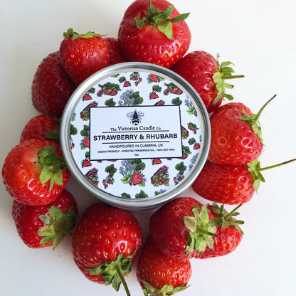 A 100ml tin of The Victorian Candle Co.'s Strawberry & Rhubarb scented candle is displayed among fresh strawberries on a white background. The recyclable tin features vibrant fruit illustrations and highlights its soy-based, vegan-friendly wax, crafted by hand in Cumbria, UK.