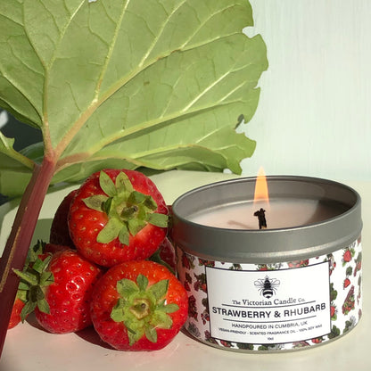 A lit Strawberry & Rhubarb Scented Candle from The Victorian Candle Co. in a recyclable 100ml tin is positioned next to fresh strawberries and a large rhubarb leaf. Bright lighting enhances the label and the vibrant red of the strawberries, while the candle's soy vegan-friendly wax ensures an eco-conscious burn.