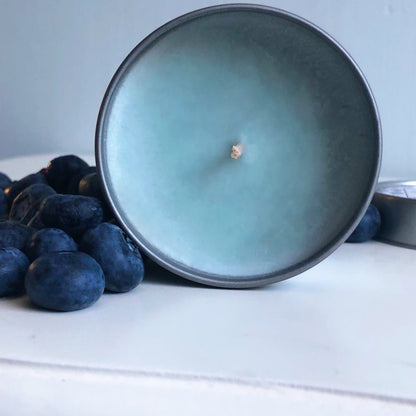 A round, blue Blueberry Pancakes-Scented Candle from The Victorian Candle Co., housed in a 100ml tin with a single wick, sits next to a small pile of blueberries on a white surface. The candle's lid is partially visible to the side, radiating the comforting aroma of blueberry pancakes.