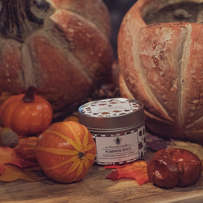 A cozy autumn scene features a Pumpkin Spice - Scented Candle in a 100ml Tin from The Victorian Candle Co., crafted from soy vegan-friendly wax and surrounded by decorative pumpkins and vibrant fall leaves. The setting exudes warmth and seasonal coziness, all packaged in recyclable tins.