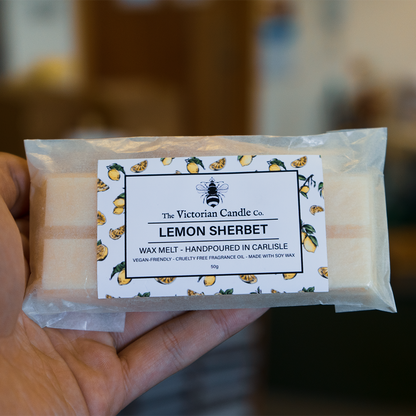 A close-up of a hand holding a wax melt package labeled Lemon Sherbet by The Victorian Candle Co. shows soy wax packaging decorated with lemon slices, highlighting its vegan-friendly and cruelty-free attributes. For lovers of fresh aromas, another delightful option is the Lime, Basil & Mandarin Wax Melt Snap Bar from The Victorian Candle Co.